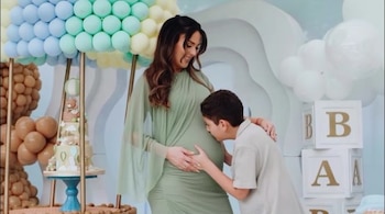 Una mujer embarazada vestida de verde sonríe mientras un niño besa su vientre en una sala decorada con globos