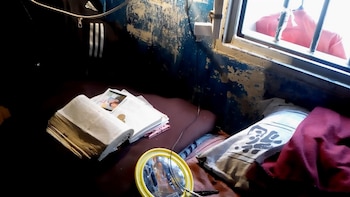 Vista de un espacio interior con pared azul descascarada, una Biblia abierta, fotos, un espejo amarillo circular y una ventana con rejas