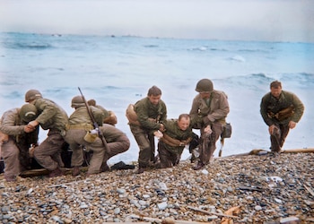 Un grupo de seis soldados en uniformes de combate y cascos, uno arrodillado siendo ayudado por otros en una playa de guijarros con el mar al fondo