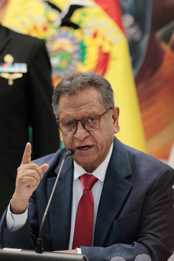 Hombre con gafas, traje oscuro y corbata roja habla en un micrófono, señalando con el índice de su mano derecha. Fondo con bandera de colores