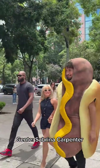 Una mujer rubia con gafas de sol camina por una calle de la ciudad junto a una persona disfrazada de hot dog, escoltada por un hombre