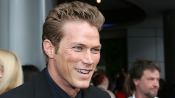 Primer plano del actor Jason Lewis sonriendo, con cabello rubio oscuro y ojos azules, vistiendo traje oscuro. Fondo borroso con otras personas