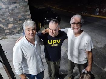 Tres hombres abrazados sobre una calle iluminada de noche junto a un vehículo oscuro