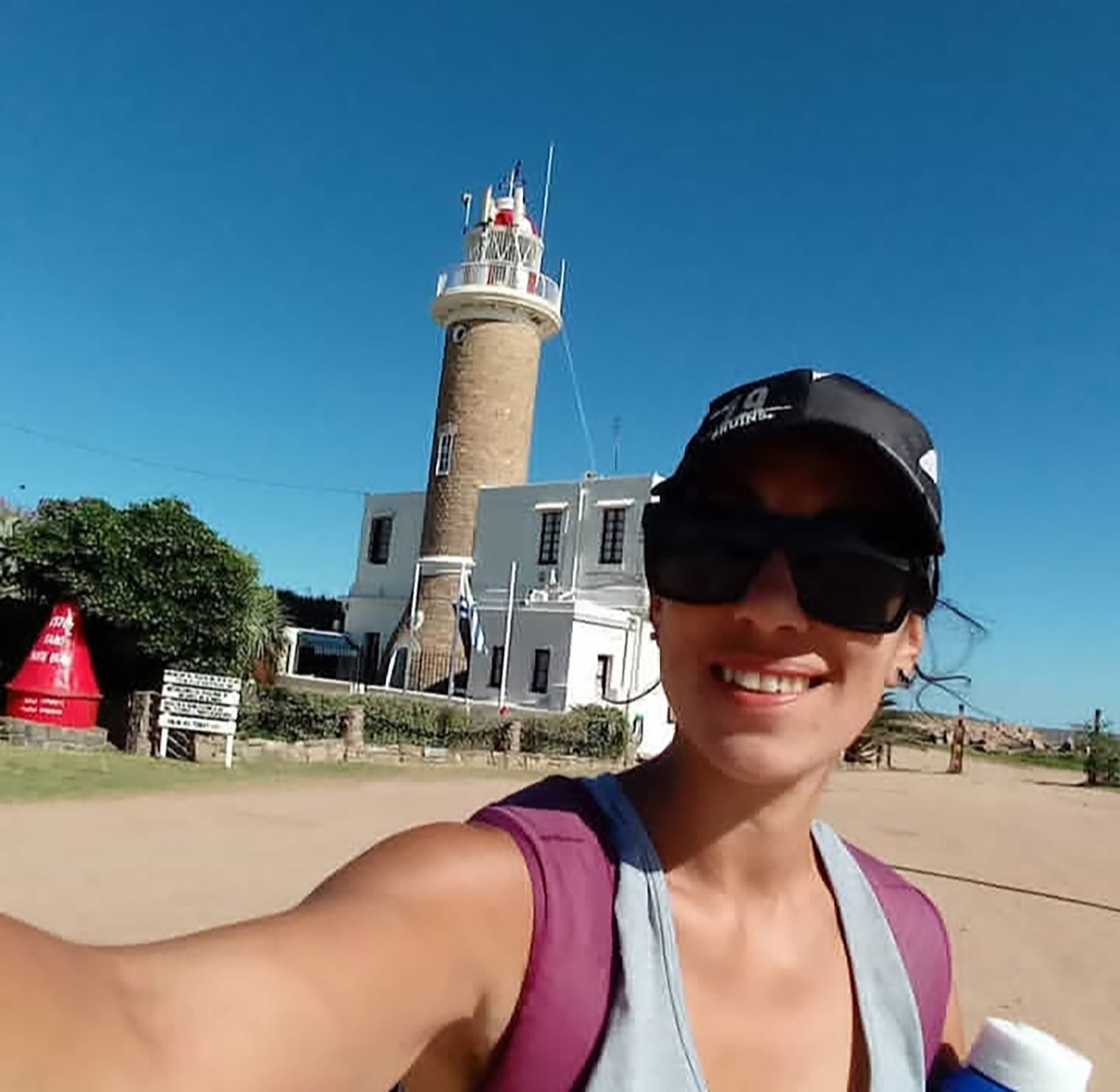 Una mujer con gafas de sol y gorra se toma una autofoto frente a un faro de piedra y una edificación blanca.