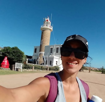 Una mujer con gorra negra y gafas de sol sonríe a la cámara en primer plano, con un faro y un edificio blanco al fondo