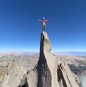 Una persona de pie en la cima de una aguja rocosa. Viste camisa rosa, pantalones verdes, mochila y gorra blanca. Al fondo hay montañas y cielo azul