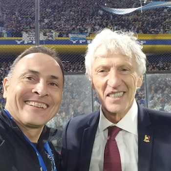 Selfie de dos hombres sonriendo en un estadio. José Pékerman, con cabello blanco, traje y corbata, está a la derecha. Detrás, gradas con aficionados