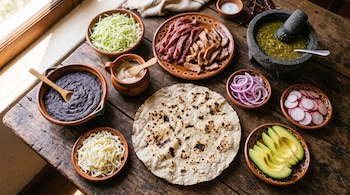Una tortilla de maíz junto a recipientes de barro con frijoles, quesillo, carne, col, cebolla, aguacate, rábanos y salsa verde en un molcajete.