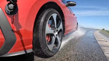 Automóvil rojo con neumático salpicando agua en pista mojada. Se observa una cámara GoPro en el lateral y cielo azul