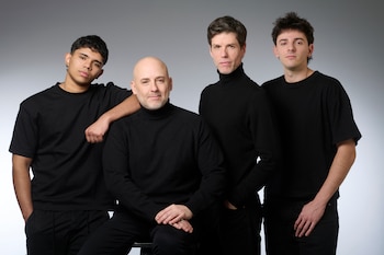 Cuatro hombres con vestimenta negra posan de frente sobre un fondo gris liso. Uno de ellos está calvo y barbado, otro tiene el cabello oscuro y usa cuello tortuga