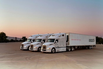 Tres camiones de carga blancos con la inscripción Waabi estacionados en fila en un estacionamiento pavimentado al atardecer