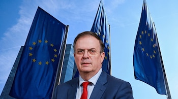Un hombre de mediana edad con gafas, traje azul marino y corbata roja, posa al frente de tres banderas de la Unión Europea bajo un cielo azul.
