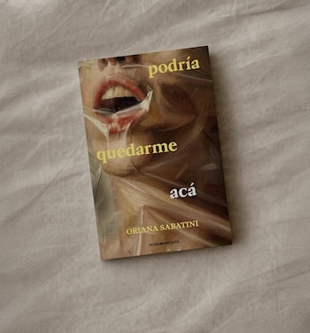 Primer plano de la portada de un libro titulado 'podría quedarme acá' de Oriana Sabatini, sobre una tela clara; muestra un rostro cubierto por plástico