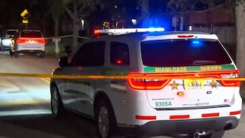 Vista trasera de un SUV blanco de la policía de Miami-Dade con luces azules y rojas encendidas, y una cinta de seguridad amarilla en primer plano