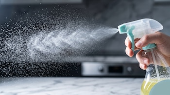 Una mano sostiene un atomizador transparente que expulsa un chorro de diminutas gotas de líquido en el aire de una cocina.