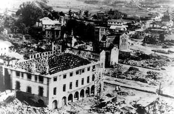 Vista aérea en blanco y negro de una ciudad en ruinas. Edificios con tejados destruidos y fachadas dañadas. Montones de escombros cubren el suelo