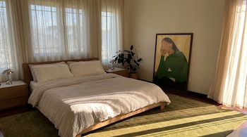 Vista de un dormitorio con cama de madera y sábanas claras, alfombra verde, mesitas de noche, una lámpara, una planta y un cuadro grande apoyado en la pared.