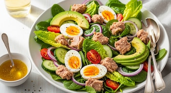 Vista aérea de una ensalada fresca en un plato blanco, con atún, aguacate, huevos duros, lechuga, espinaca, tomate, pepino y cebolla morada.