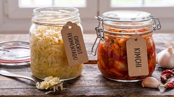 Dos frascos de vidrio con chucrut y kimchi etiquetados sobre una mesa de madera junto a un tenedor, un diente de ajo y pimientos secos.