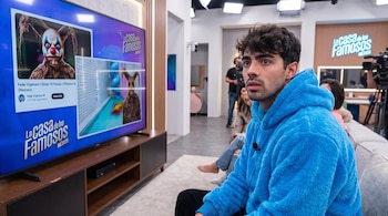 Un hombre joven con sudadera azul mira un televisor con la miniatura de un payaso y el logo de La Casa de los Famosos México. Hay cámaras en el fondo.
