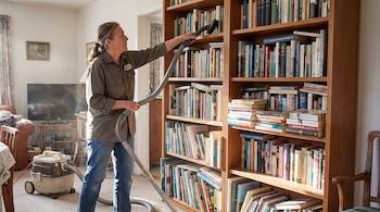 Una mujer de cabello recogido, camisa verde oliva y jeans aspira un librero de madera con estantes repletos de libros. Una aspiradora beige está en el suelo.