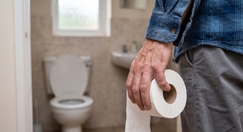 Primer plano de la mano de un hombre con camisa a cuadros que sostiene un rollo de papel higiénico con un inodoro desenfocado al fondo