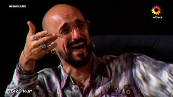 Primer plano de un hombre calvo y barbudo con gafas y camisa estampada, riendo a carcajadas. Se aprecian anillos en sus dedos y tatuajes en su mano