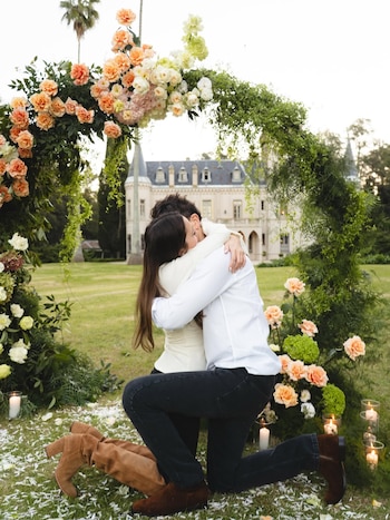 Un hombre de rodillas abraza a una mujer bajo un arco de flores en un jardín con césped. Alrededor hay pétalos blancos y velas. De fondo, un castillo