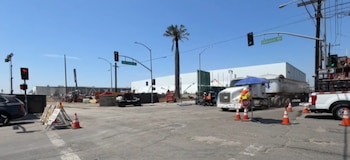 Intersección de calles con semáforos, señalización vial, obreros, vehículos de construcción, un camión y conos de tráfico bajo un cielo azul