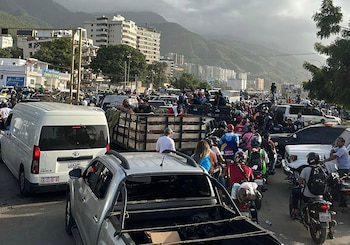 Calle concurrida con furgonetas, camiones, motocicletas y coches, cargados de personas y equipo, y edificios frente a montañas bajo un cielo nublado