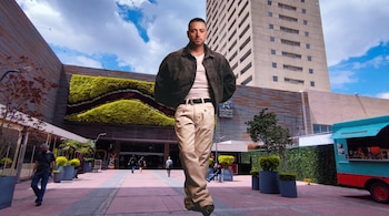 Un hombre de pie viste chaqueta oscura y pantalones claros frente a un edificio con jardín vertical y una plaza urbana.