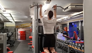Vista trasera de un hombre con camiseta blanca y pantalón corto negro realizando dominadas asistidas en una máquina de gimnasio