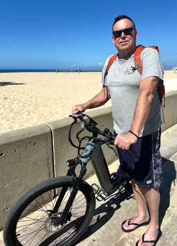 Hombre con gafas de sol, camiseta gris, shorts, sandalias y mochila naranja posa junto a bicicleta híbrida gris en paseo marítimo con playa y mar