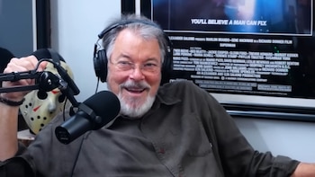 Jonathan Frakes, un hombre con barba gris y gafas, sonríe a la cámara con auriculares y un micrófono. Detrás de él, una máscara de Jason y un póster de Superman