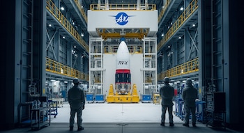 Interior de hangar. Cohete RV-X blanco, rojo y negro con logo JAXA en plataforma de pruebas. Tres personas con casco y uniforme observan la escena.