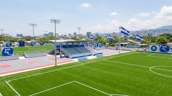 Complejo deportivo con cancha de fútbol de césped sintético y cancha de baloncesto, gradas grises, postes de luz LED, logos azules del BCIE y banderas de El Salvador.