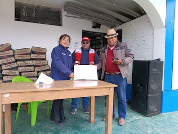 Tres personas: mujer de chaqueta azul, hombre de chaleco rojo y gorra, hombre de sombrero. Frente a una mesa de madera. Al fondo, bolsas de cemento apiladas