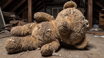Peluche de oso marrón sucio y roto sobre un suelo de madera con residuos. Al fondo se observan vigas de madera y objetos.