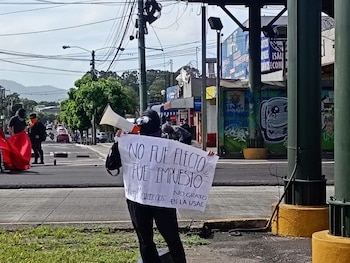 Un manifestante sostiene pancarta que dice 'NO FUE ELECTO FUE IMPUESTO' y un megáfono; otras personas y una calle con vehículos
