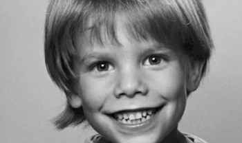 Primer plano en blanco y negro del rostro de un niño pequeño sonriendo, con el cabello claro y corto, mirando directamente a la cámara. Sus dientes son visibles