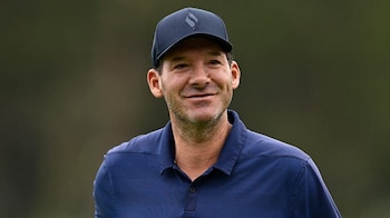 Primer plano de un hombre sonriente, vestido con una camiseta polo azul marino y una gorra azul oscura, sobre un fondo verde desenfocado