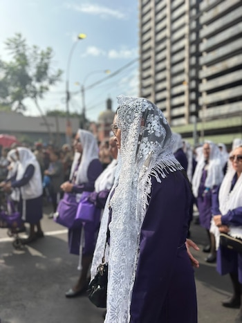 Mujeres vestidas con túnicas moradas y mantillas blancas de encaje permanecen de pie en una calle urbana junto a un edificio alto