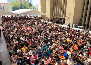 Una multitud de personas sentadas en hileras de sillas al aire libre junto a un edificio de concreto y bajo una carpa blanca
