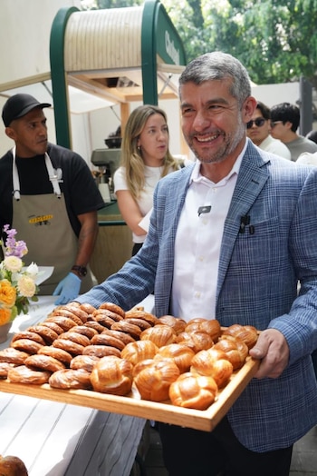 Mauricio Tabe sonríe mientras sostiene una bandeja de madera con pan dulce variado y croissants. Detrás, se ven personas y un puesto de venta