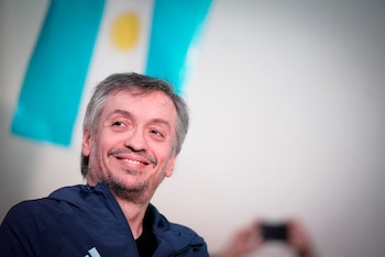 Primer plano de Máximo Kirchner, hombre con barba, sonriendo con una campera azul, y una bandera de Argentina en el fondo