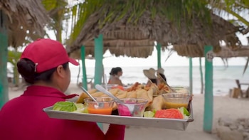Persona de espalda con gorra roja y camisa roja lleva bandeja metálica con comida frita, salsas, limas, aguacate y repollo en una playa