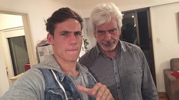 Un hombre joven con sudadera y chaleco de mezclilla, señalando, junto a un hombre mayor de pelo blanco y camisa a cuadros, posan en el interior de una casa.