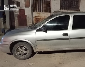 Un coche plateado, ligeramente sucio, está estacionado de noche frente a un edificio con una reja y ventanas. El logotipo del MPA de Jujuy es visible