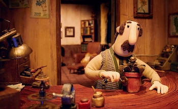 Personaje animado stop-motion con bigote, suéter y chaleco, en un escritorio de madera. Manipula una planta en maceta roja, con lámpara y herramientas alrededor
