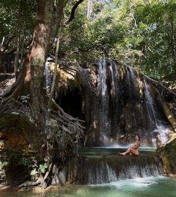 Una mujer con traje de baño blanco está sentada al borde de un estanque natural, frente a una cascada rodeada de vegetación